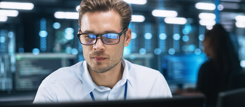 Young man wearing glass sitting in datacenter looking at monitors