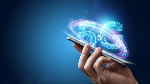 Hand holding mobile phone with 5G text hovering on blue background